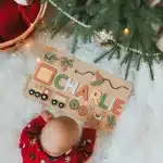 A baby lies next to a Christmas tree with a wooden sign.