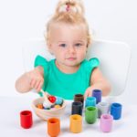 Ein kleines Mädchen spielt mit Montessori Holzspielzeug Kugel und Sortierbecher und sortiert bunte Kugeln und Becher.