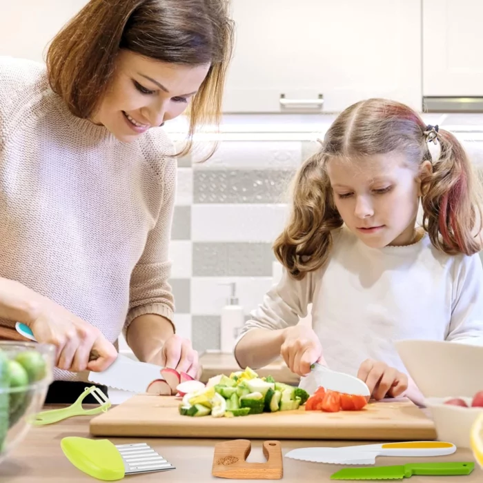 Un adulte et un enfant coupent ensemble des légumes dans la cuisine. Divers ustensiles de cuisine et couteaux, dont le "Couteau Enfant - Ensemble de Cuisine 13 pièces - dès 2 ans" aux couleurs vives, sont placés sur une planche à découper en bois devant eux.