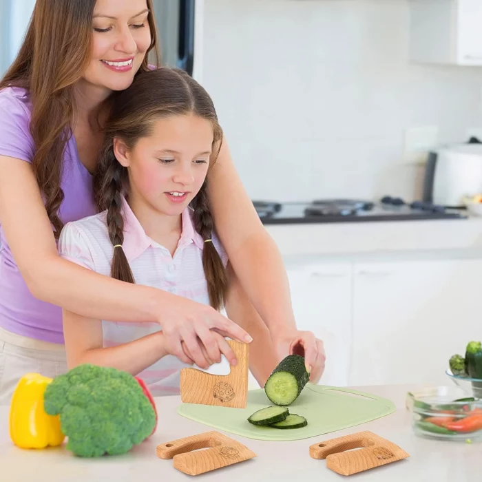 Un adulte et un enfant coupent des concombres sur une planche à découper verte dans une cuisine, en utilisant le Couteau Enfant de l'Ensemble de Cuisine 13 pièces - dès 2 ans, avec du poivron et du brocoli à proximité. Trois ustensiles de cuisine en bois sont posés sur le comptoir.