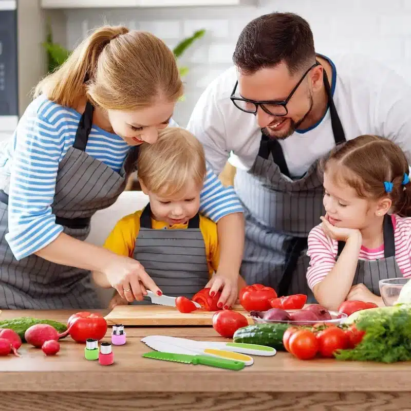 Une famille de quatre personnes, tous vêtus de tabliers rayés, est réunie autour d'une table de cuisine. Les parents aident leurs deux jeunes enfants à couper des légumes, dont des tomates et des poivrons, avec le « Couteau Enfant - Ensemble de Cuisine 13 pièces - dès 2 ans », conçu pour une utilisation sécuritaire par les petites mains.