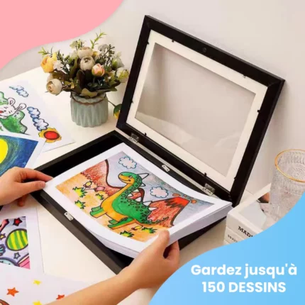 Sur un bureau, une personne place un dessin de dinosaure format A4 dans le Cadre pour dessins d'enfant noir, entouré d'autres œuvres, d'un vase de fleurs et d'un verre. Texte : "Stockez et exposez jusqu'à 150 dessins au format A4.