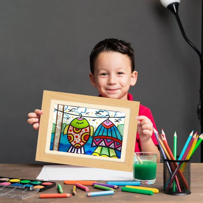 Un enfant souriant affichant un Cadre pour dessins d’enfants coloré - stockez et exposez 150 dessins - dessin encadré taille A4 avec des fournitures artistiques autour.