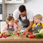 Un homme avec deux enfants préparant de la nourriture dans une cuisine, entouré de divers légumes frais et de cadres dessinant.