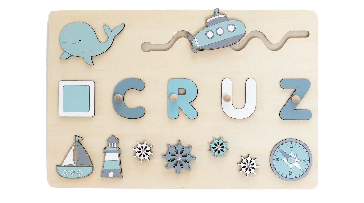 Planche en bois portant le nom « Cruz » avec des découpes décoratives sur le thème nautique et maritime, notamment une baleine, un sous-marin, un voilier, un phare, une boussole et des jeux de réflexion inspirés des casse-tête.