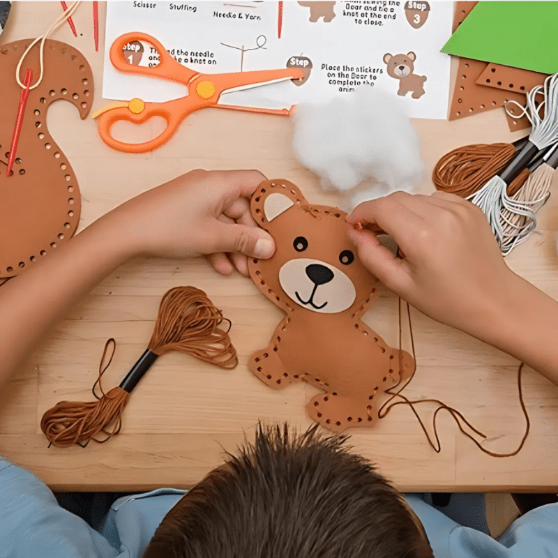 Un enfant coud joyeusement un ours en peluche avec du fil marron sur une table en bois, entouré de ciseaux, d'instructions et de fils à broder colorés de son Kit de Couture Enfants - Porte-clés à Faire Soi-Même Sans se piquer le doigt.