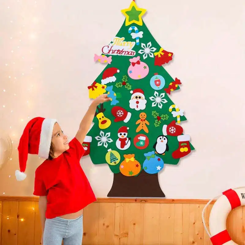 Un enfant portant un chapeau de Père Noël décore un sapin de Noël en feutre (Sapin Noël) sur le mur avec divers ornements, notamment des flocons de neige, des cannes à sucre et une étoile.