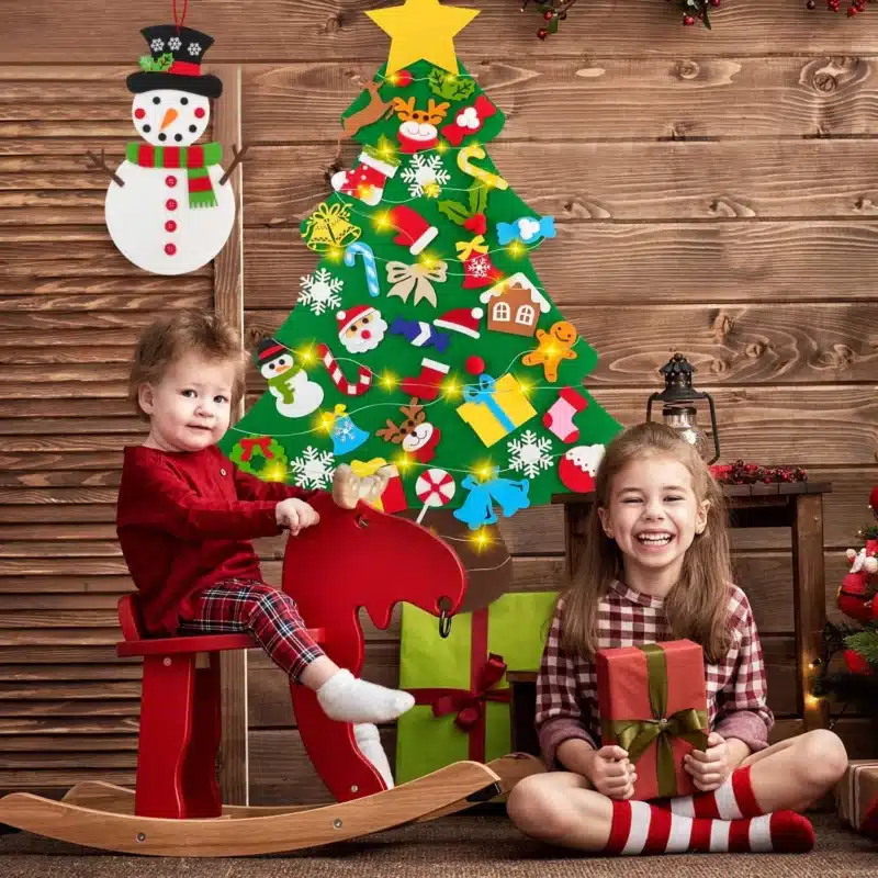 Deux enfants en tenues de fête, l'un assis sur un cheval à bascule et l'autre tenant un cadeau, avec un sapin en feutre décoré en arrière-plan sur un mur en bois, capturant la magie de Noël.