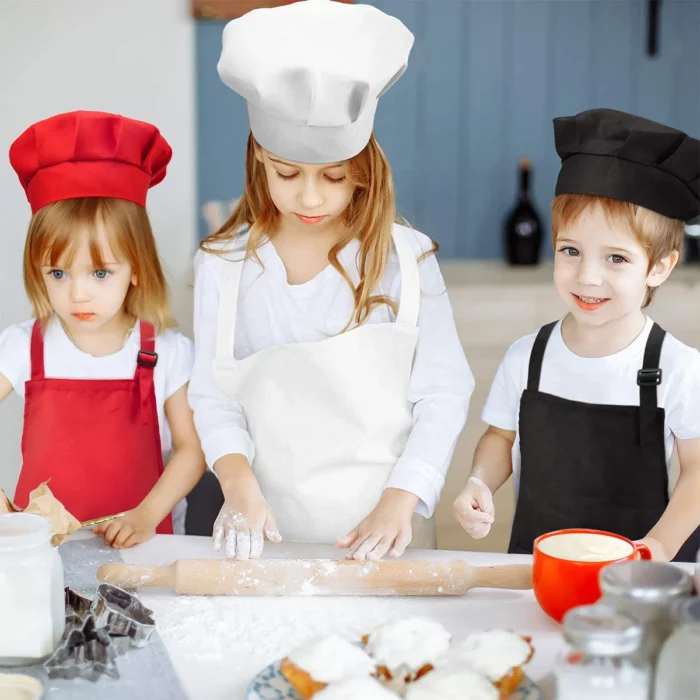 Trois enfants portant des toques et des tabliers de chef cuisinent ensemble, étalent la pâte et sont entourés d'ingrédients de pâtisserie sur la table.