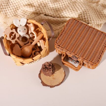 Un panier en osier avec des jouets en bois, comme un lapin et des hochets, est posé à côté d'un Coffret naissance hochets bois en osier fermé sur une surface crème avec une couverture texturée.