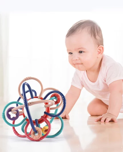 Un bébé en rose rampe sur un sol léger vers le jouet multifonction Hochet- Montessori aux boucles colorées entrelacées, découvrant la joie du jeu multifonction.