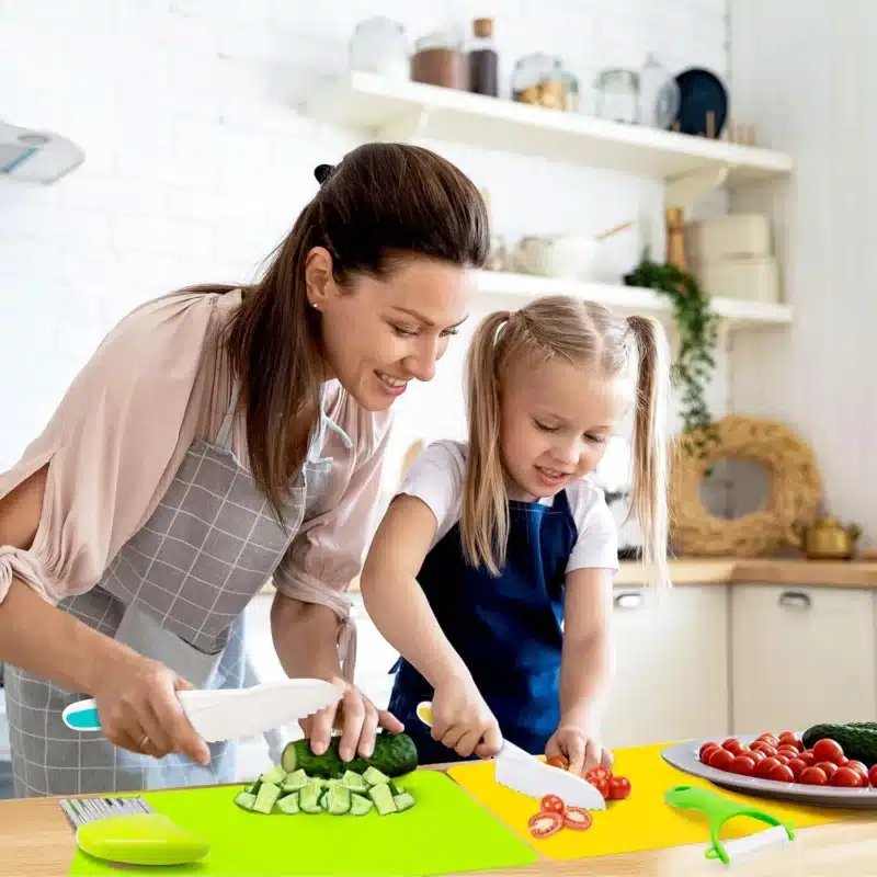 Dans une cuisine animée rappelant une page d’atterrissage d’Elementor, une femme et une jeune fille coupent joyeusement des légumes. La femme coupe habilement des concombres tandis que la fille tranche des tomates, toutes deux brandissant des couteaux en plastique au-dessus de planches à découper colorées qui semblent mettre en valeur leur aventure culinaire commune.