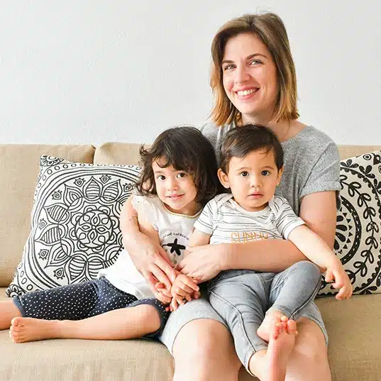 Une femme souriante est assise sur un canapé avec deux jeunes enfants, un de chaque côté, qui partagent des histoires. Ils sont dans un salon confortable avec des coussins à motifs et un Couteau Enfant bien en sécurité en arrière-plan.