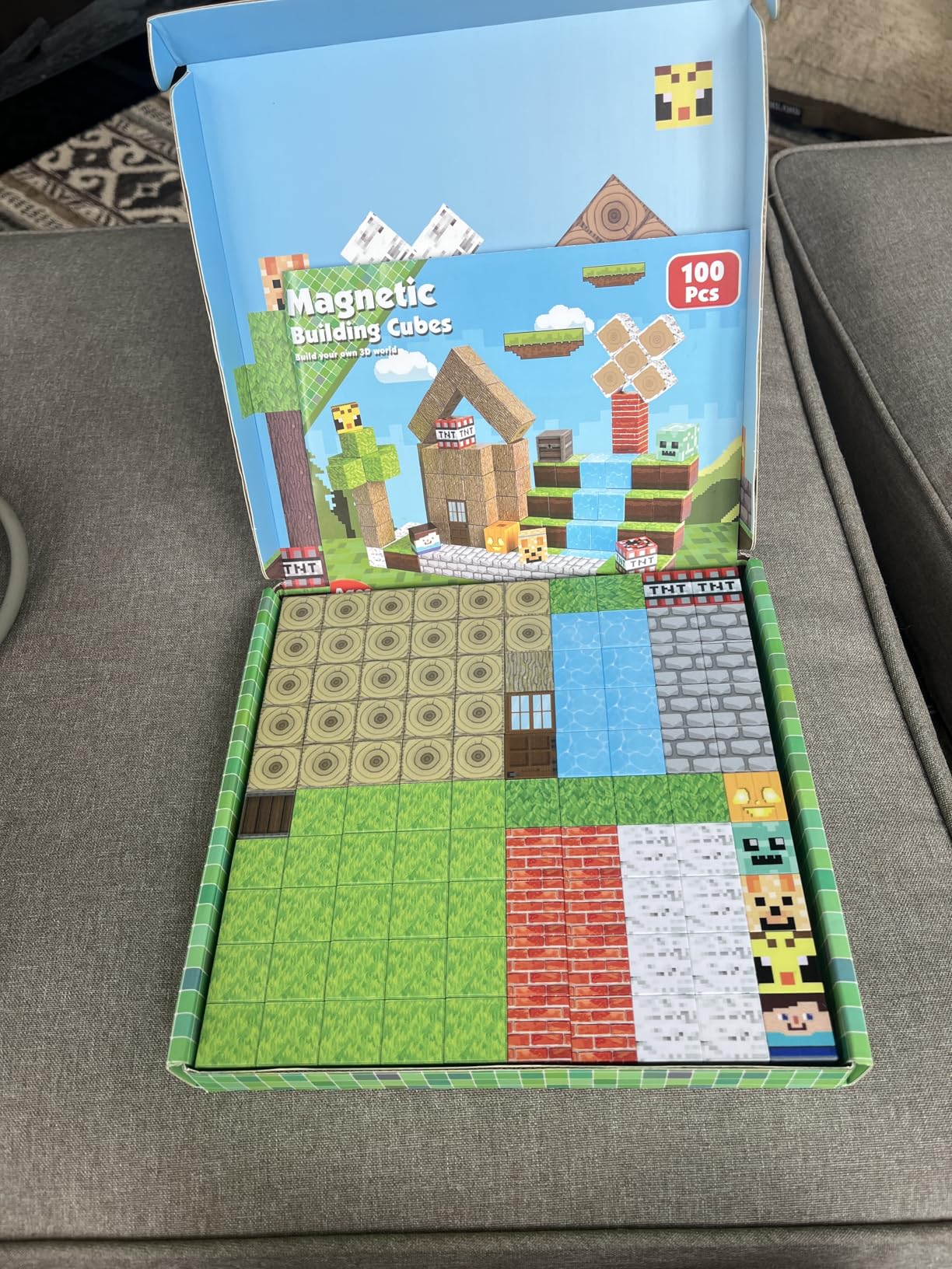 Une boîte de « cubes de construction magnétiques » contenant 100 pièces est ouverte, montrant des tuiles à motifs de style Minecraft disposées soigneusement à l'intérieur.